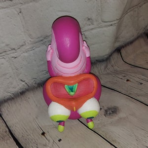 Op de afbeelding: Een roze en oranje figuur met een groot hoofd en kleine benen. Het figuur heeft een groot, rond hoofd met een roze gezicht en een kleine, oranje mond. Het lichaam van het figuur is roze en heeft een groot, oranje hartvormig ontwerp aan de voorkant. De benen van het figuur zijn klein en wit met groene en gele accenten.