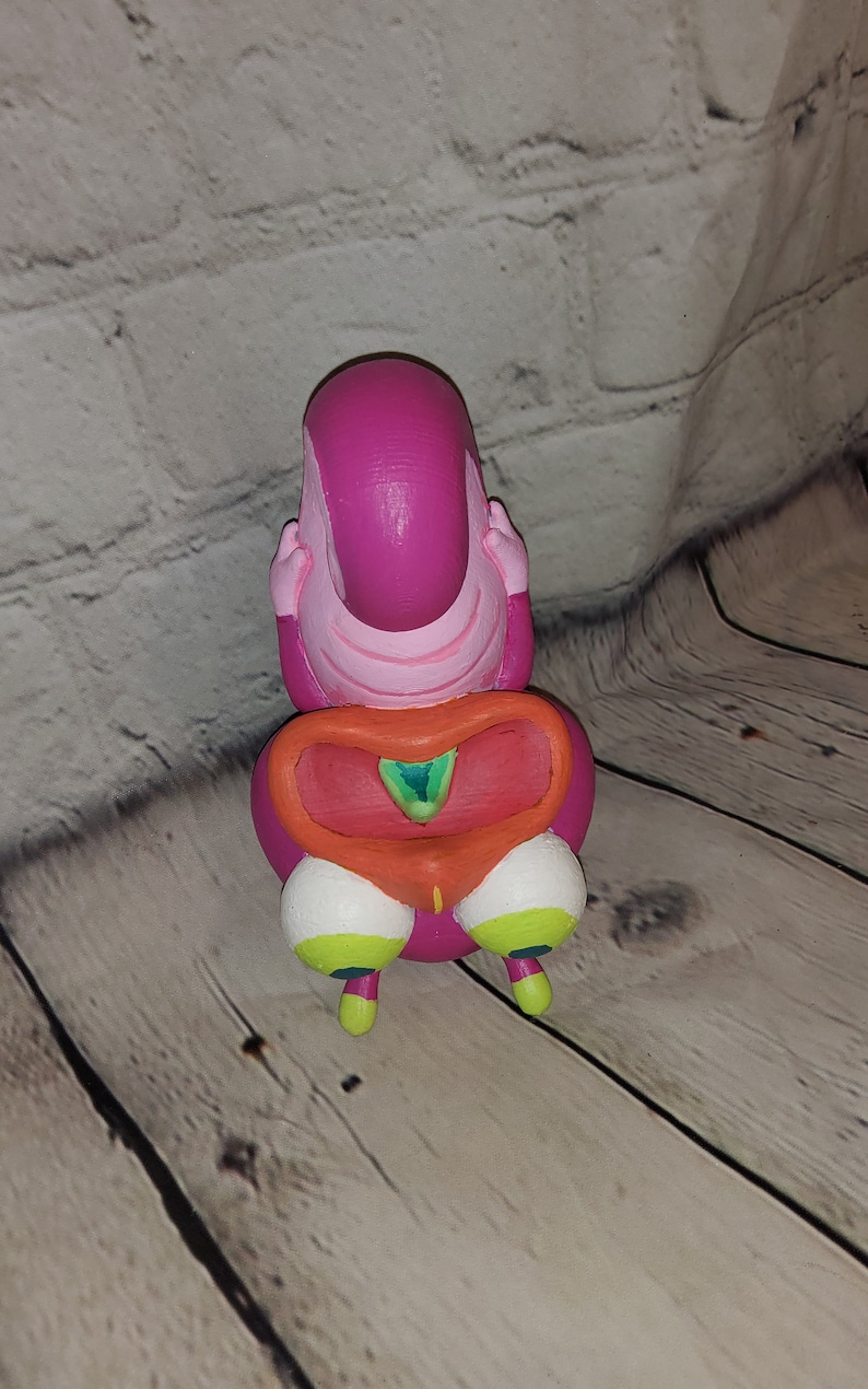 Op de afbeelding: Een roze en oranje figuur met een groot hoofd en kleine benen. Het figuur heeft een groot, rond hoofd met een roze gezicht en een kleine, oranje mond. Het lichaam van het figuur is roze en heeft een groot, oranje hartvormig ontwerp aan de voorkant. De benen van het figuur zijn klein en wit met groene en gele accenten.