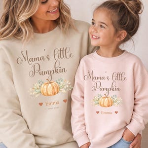 Könnte beinhalten: Ein beigefarbener Pullover und ein hellrosa Pullover, beide mit dem Schriftzug "Mama's little Pumpkin" in brauner Schreibschrift, einer Aquarell-Kürbisillustration mit grünen Blättern sowie dem Namen "Emma" mit kleinen Herzen und "since 2026".