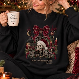 Könnte beinhalten: Ein schwarzes Langarm-Sweatshirt mit einem Grafikdruck eines Totenkopfs mit Weihnachtsmütze, einer großen roten Schleife, Büchern, Kerzen und einem Raben. Der Text lautet Merry Gothmas, Julia's Gothmas Club und Est 2026 in dekorativer Schrift.