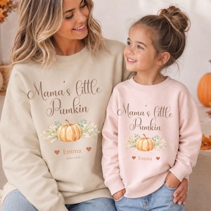 Könnte beinhalten: Ein beigefarbenes Sweatshirt und ein hellrosa Sweatshirt, beide mit dem Text Mama's little Pumkin, einer Aquarell-Kürbisillustration mit Blattwerk sowie dem Namen Emma und Herzsymbolen. Das beige Sweatshirt enthält zusätzlich den Text since 2021.