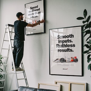 May include: Two framed posters with black frames are displayed on a white wall. The top poster reads "Speed mattered more than sense." The bottom poster says "Smooth inputs. Ruthless results." A person is on a ladder.