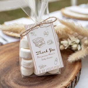 May include: A clear cellophane bag filled with white marshmallows, tied with twine, and a decorative tag. The tag has a striped border and reads "Thank You For joining Olivia's baby shower" with baby-themed illustrations.