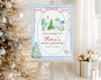May include: A rectangular sign on a wooden easel featuring a light blue gingham border, a red bow at the top, and a central oval frame. It displays a watercolor village, a decorated Christmas tree, a deer, and the text WELCOME TO Olivia's BABY SHOWER AUGUST 10, 20XX.