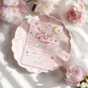May include: A pink baby shower invitation with a teddy bear on a cloud design. The invitation features gold stars, a crescent moon, and the text "We're on Cloud Nine." The invitation is on a pink plate with pink flowers.