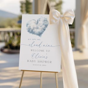 以下が含まれることがあります： 金色のイーゼルに立てられた白いベビーシャワーのウェルカムサイン。青い雲のハートのグラフィックと「we are on cloud nine, Welcome to Olivia's Baby Shower, August 10, 20XX」の文字が特徴です。クリーム色のリボンと布が側面を覆っています。