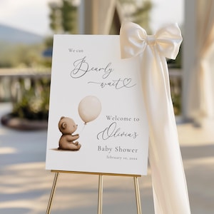 May include: A baby shower welcome sign on a gold easel. The sign features a watercolour illustration of a teddy bear holding a balloon. Text reads "We can Bearly wait" and "Welcome to Olivia's Baby Shower February 10, 20xx."