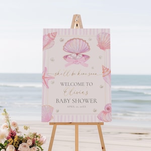 May include: A baby shower welcome sign on an easel. The sign features a seashell theme with pink and gold accents. Text reads "shell be here soon", "Welcome to Olivia's Baby Shower" and the date.