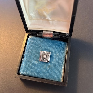 May include: A small, square silver-toned pin with an ornate design, set on a blue velvet cushion inside a black box. The box is open, revealing the pin's intricate details and the contrasting textures of the velvet and the shiny pin.