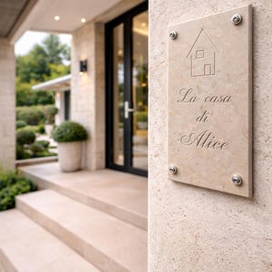 Puede incluir: Placa rectangular de piedra beige con las palabras "La casa di Alice" grabadas en cursiva. La placa está montada en una pared de piedra texturizada con cuatro tornillos plateados. Un simple contorno de casa está encima del texto.