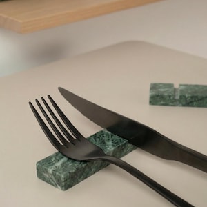 Può includere: Set di forchetta e coltello neri appoggiati su supporti in marmo verde. La forchetta ha quattro rebbi e il coltello una lama seghettata. Il set è presentato su una superficie chiara, ideale per la tavola.