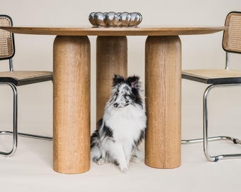 May include: A round wooden dining table with three cylindrical legs, flanked by two chairs with woven backs and seats. A small, silver decorative bowl sits on the table. A dog sits under the table.