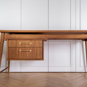May include: A mid-century modern wooden desk with two drawers and brass handles. The desk has a long, rectangular top and angled legs. The wood has a warm, natural finish. The desk is set against a white background.