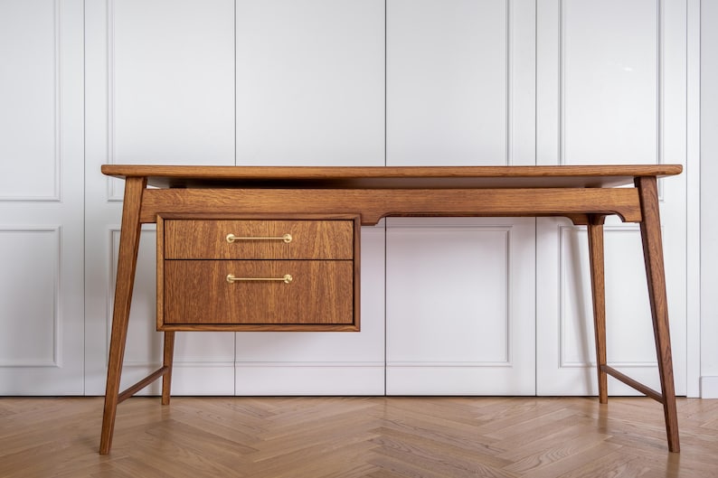 May include: A mid-century modern wooden desk with two drawers and brass handles. The desk has a long, rectangular top and angled legs. The wood has a warm, natural finish. The desk is set against a white background.
