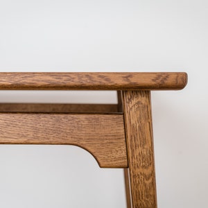 May include: Close-up of a wooden table with a natural finish. The table has a rectangular top and a lower shelf, supported by four legs. The wood grain is visible, and the table is set against a white background.