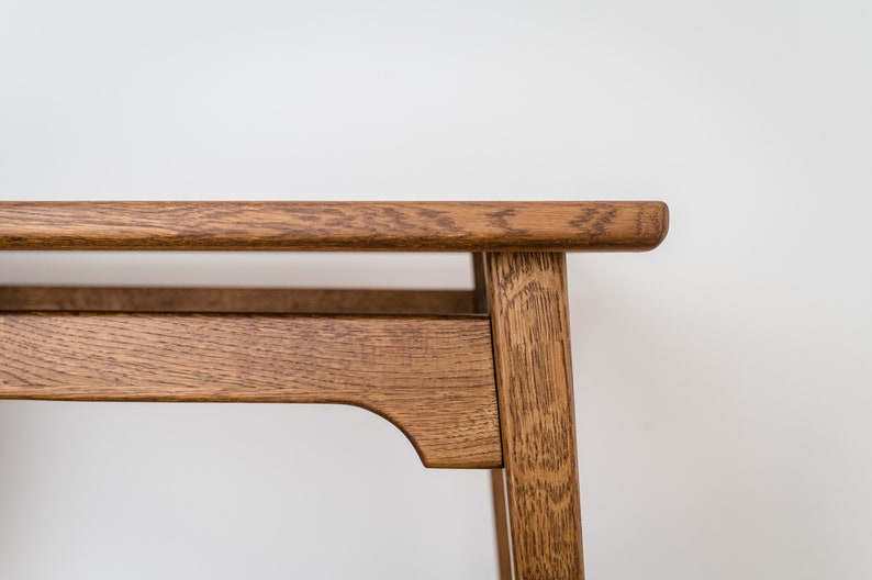 May include: Close-up of a wooden table with a natural finish. The table has a rectangular top and a lower shelf, supported by four legs. The wood grain is visible, and the table is set against a white background.