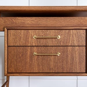 May include: Close-up of a wooden desk with two drawers. The desk is made of a medium-toned wood with visible grain. The drawers have brass bar pulls. The desk has a minimalist design with angled legs.