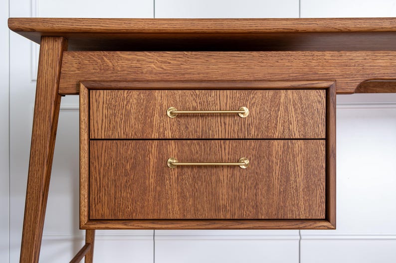 May include: Close-up of a wooden desk with two drawers. The desk is made of a medium-toned wood with visible grain. The drawers have brass bar pulls. The desk has a minimalist design with angled legs.