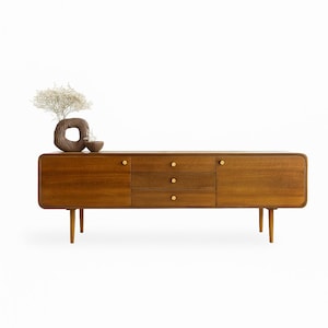 May include: A mid-century modern style wooden sideboard with three drawers and two cabinet doors. The cabinet has brass knobs and is supported by four tapered legs. A decorative vase and bowl sit on top.