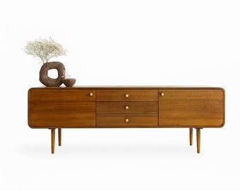 May include: A mid-century modern style wooden sideboard with three drawers and two cabinet doors. The cabinet has brass knobs and is supported by four tapered legs. A decorative vase and bowl sit on top.