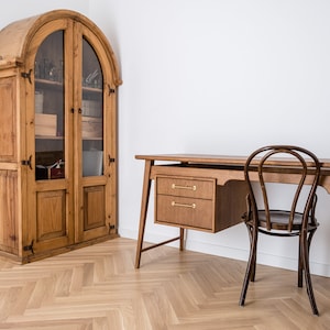May include: A wooden desk with two drawers and a matching chair. A tall, antique wooden cabinet with glass doors stands to the left. The room has a light wood floor and white walls.