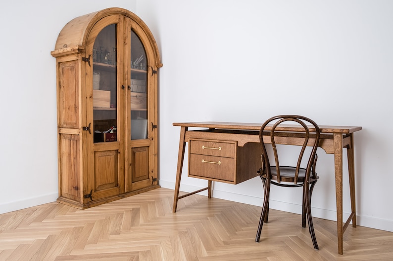 May include: A wooden desk with two drawers and a matching chair. A tall, antique wooden cabinet with glass doors stands to the left. The room has a light wood floor and white walls.
