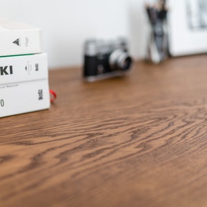 May include: A stack of three white books with black text on a wooden surface. The top book reads "LK HOME", the middle book reads "SZEWSKI", and the bottom book reads "SZTUKA POLSKIEGO PLAKATU". A vintage camera is in the background.