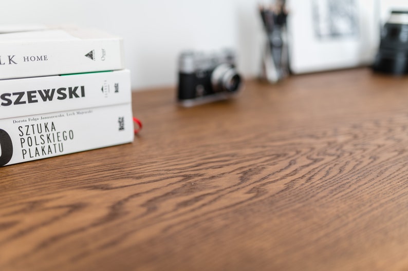 May include: A stack of three white books with black text on a wooden surface. The top book reads "LK HOME", the middle book reads "SZEWSKI", and the bottom book reads "SZTUKA POLSKIEGO PLAKATU". A vintage camera is in the background.