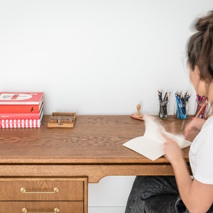May include: A wooden desk with a stack of red books, a wooden stamp set, and pencils in glass jars. A person is seated at the desk, working on a notebook. The desk has a drawer with gold-colored handles.