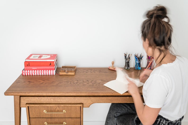 May include: A wooden desk with a stack of red books, a wooden stamp set, and pencils in glass jars. A person is seated at the desk, working on a notebook. The desk has a drawer with gold-colored handles.