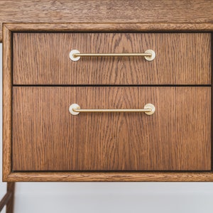 May include: Close-up of a wooden desk with two drawers. The drawers are made of dark brown wood with a visible grain pattern. Each drawer has a gold-colored, horizontal bar handle. The desk appears to be part of a larger furniture set.