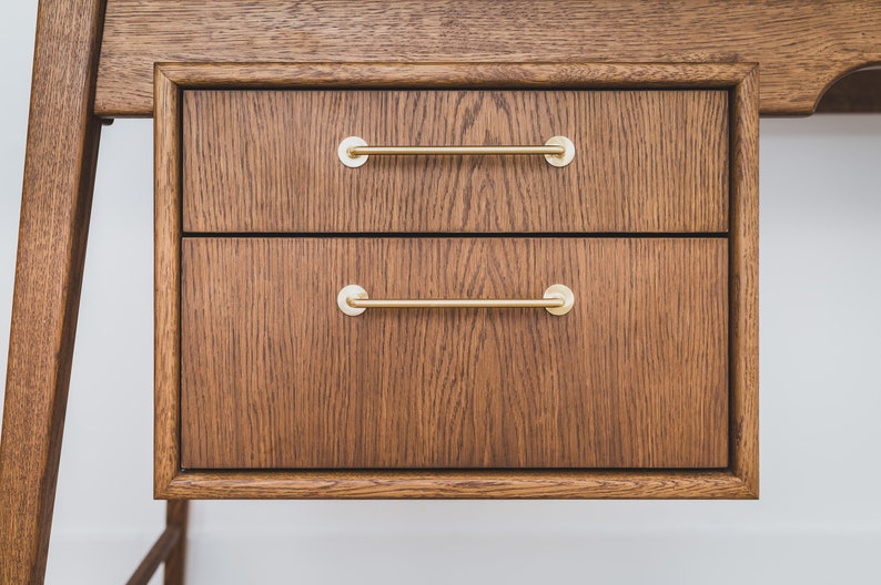May include: Close-up of a wooden desk with two drawers. The drawers are made of dark brown wood with a visible grain pattern. Each drawer has a gold-colored, horizontal bar handle. The desk appears to be part of a larger furniture set.