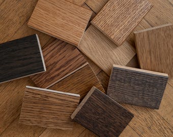 May include: A collection of wooden flooring samples in various shades of brown, from light oak to dark walnut. The samples are rectangular and show different wood grain patterns, arranged on a wooden floor.