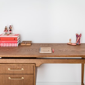 May include: A wooden desk with two drawers, a stack of red books, and a small notebook. A pencil holder with colored pencils and a small wooden box are also on the desk. Two figurines stand atop the books. The desk is made of wood.
