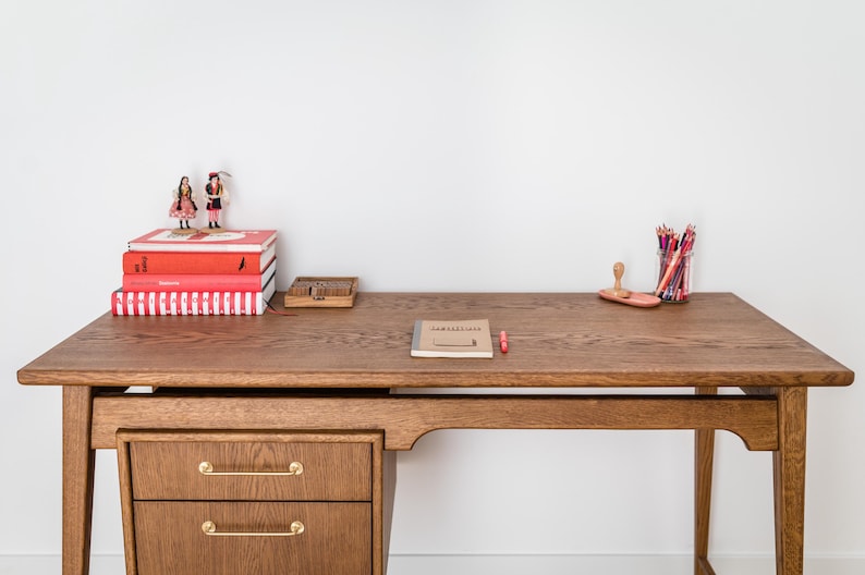 May include: A wooden desk with two drawers, a stack of red books, and a small notebook. A pencil holder with colored pencils and a small wooden box are also on the desk. Two figurines stand atop the books. The desk is made of wood.