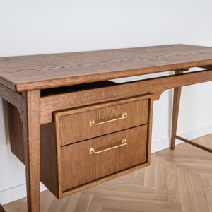 May include: A wooden desk with a warm brown finish. It features a rectangular tabletop, two drawers with gold-colored handles, and slender legs. The desk is designed with a mid-century modern aesthetic, suitable for a home office or workspace.