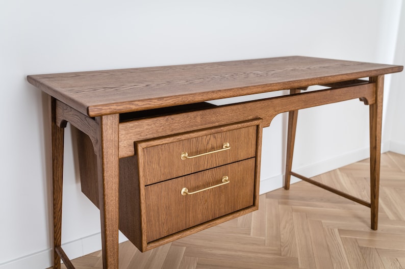 May include: A wooden desk with a warm brown finish. It features a rectangular tabletop, two drawers with gold-colored handles, and slender legs. The desk is designed with a mid-century modern aesthetic, suitable for a home office or workspace.