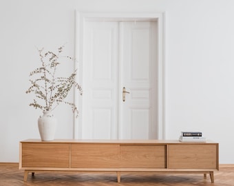 May include: A light oak wood media console with three sections and a mid-century modern design. A white vase with dried branches sits on the left side. A stack of books rests on the right side. The console is in front of a white door.