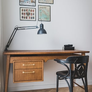 May include: A wooden desk with two drawers and brass handles, a black desk lamp, and a black chair. The wall features framed prints, including a New Yorker cover and maps. The desk is set on a herringbone wood floor.