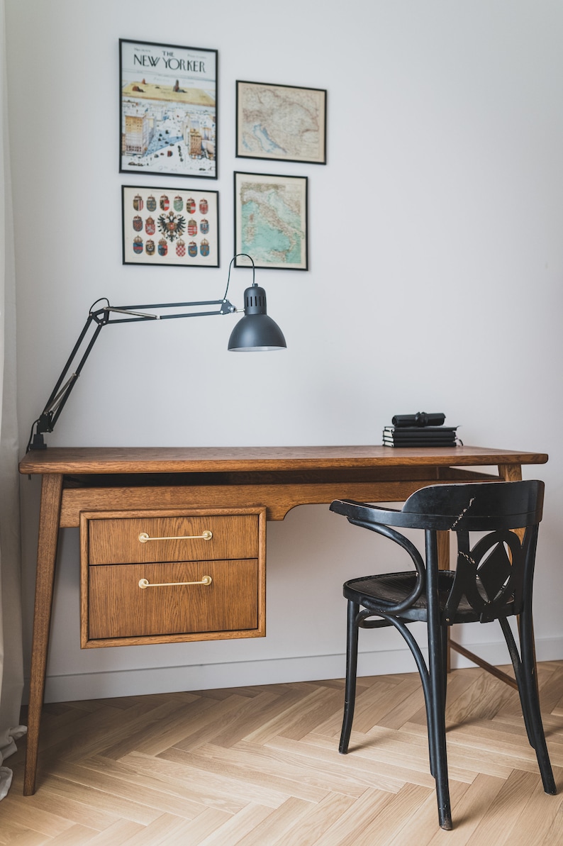May include: A wooden desk with two drawers and brass handles, a black desk lamp, and a black chair. The wall features framed prints, including a New Yorker cover and maps. The desk is set on a herringbone wood floor.