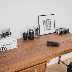 May include: A wooden desk with various items, including two cameras, a framed drawing, a stack of notebooks, and a black pencil case. Books are stacked on the left side. The desk has a warm, natural wood tone.