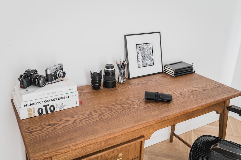 May include: A wooden desk with various items, including two cameras, a framed drawing, a stack of notebooks, and a black pencil case. Books are stacked on the left side. The desk has a warm, natural wood tone.