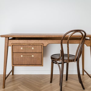May include: A wooden desk with two drawers and brass handles, paired with a curved-back wooden chair. The desk has a natural wood finish and a minimalist design, suitable for a home office or workspace. The chair has a dark brown finish.