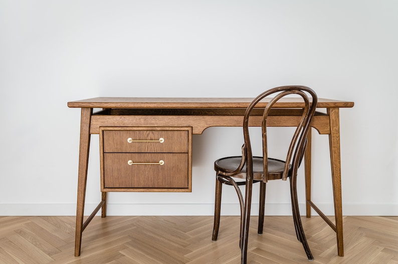 May include: A wooden desk with two drawers and brass handles, paired with a curved-back wooden chair. The desk has a natural wood finish and a minimalist design, suitable for a home office or workspace. The chair has a dark brown finish.