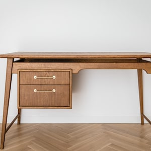 May include: A wooden desk with a warm brown finish. It features two drawers with brass handles on the left side and a spacious tabletop. The desk has a mid-century modern design with angled legs, suitable for a home office or workspace.