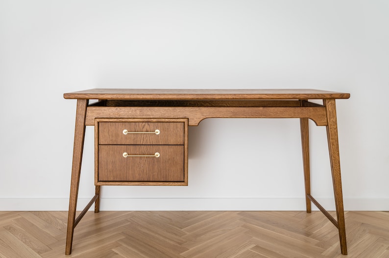 May include: A wooden desk with a warm brown finish. It features two drawers with brass handles on the left side and a spacious tabletop. The desk has a mid-century modern design with angled legs, suitable for a home office or workspace.