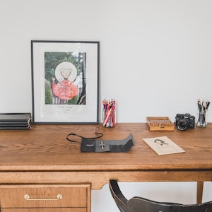 May include: A wooden desk with various items, including a framed artwork, a stack of black notebooks, colored pencils, a camera, and a pencil case. The desk has a drawer with a brass handle and a black chair is tucked underneath.