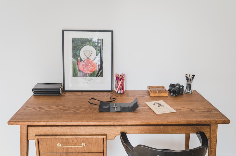 May include: A wooden desk with various items, including a framed artwork, a stack of black notebooks, colored pencils, a camera, and a pencil case. The desk has a drawer with a brass handle and a black chair is tucked underneath.