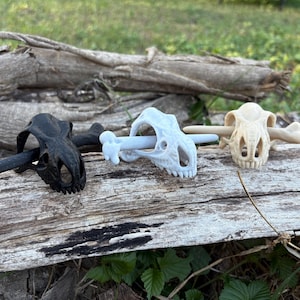 May include: Three dinosaur skull-shaped bottle stoppers in black, light blue, and beige. Each stopper has a bone-shaped handle. The stoppers are displayed on a weathered wooden log, with green foliage in the background.