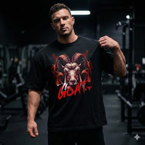 May include: Black t-shirt featuring a detailed illustration of a ram's head with fiery accents and the text "The GOAT" in red. The graphic design is centered on the front of the shirt.